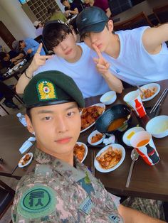 three young men sitting at a table with plates of food in front of them and pointing to the camera