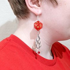 If you want to make a statement, and that statement is FIRE, these earrings are for you. A bright red and black D20 and D8 hang from a silver-plated ear hook. Below it hangs a gorgeous flame charm and a round red crystal. Great earrings for a fire domain cleric, or any fire-wielding character. These are perfect for players of D&D, Pathfinder, and other tabletop RPGs -- or for anyone who wants to geek out with a fun accessory. The flame itself is about 1-1/2 inches long, and the earrings hang nea Fire Earrings, Geeky Jewellery, Dice Earrings, Fire Jewelry, D20 Dice, Unusual Earrings, Geek Out, Drawstring Pouch, Red Crystals