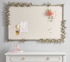 a white dresser with flowers on it and a mirror hanging above the top that is covered in gold leaf trim