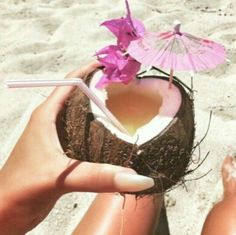 a person is holding a coconut drink in their hand on the beach with an umbrella