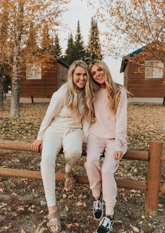 two women sitting on a wooden fence in front of a cabin and trees with leaves all over the ground