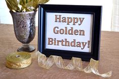 a happy golden birthday sign next to a roll of gold ribbon and a vase with flowers