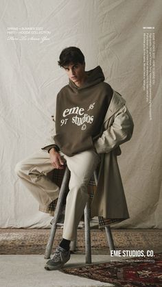 a young man sitting on top of a chair in front of a white wall wearing a hoodie