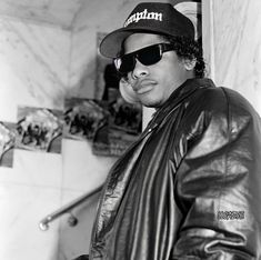 a black and white photo of a man wearing sunglasses, a leather jacket, and a hat