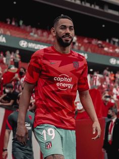 a man in red shirt and green shorts on soccer field with crowd watching from bleachers