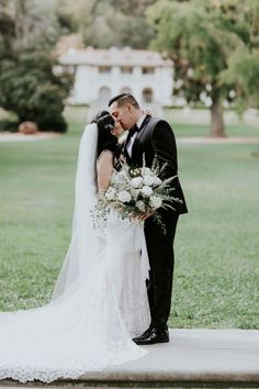 a bride and groom kissing in front of a large white building with greenery on the lawn