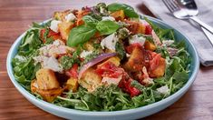 a blue bowl filled with salad on top of a wooden table next to a fork and knife