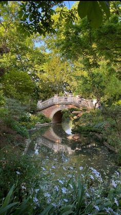 a bridge over a small river surrounded by trees and flowers with people walking on it