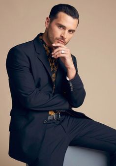 a man sitting on top of a chair wearing a black suit and yellow shirt with his hand under his chin