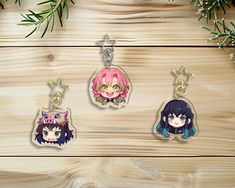 three keychains with anime characters on them sitting on a wooden table next to pine branches