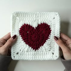 two hands holding up a crocheted square with a red heart on the front