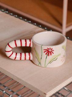 a white cup sitting on top of a wooden table next to a wire rack with a red and white striped handle