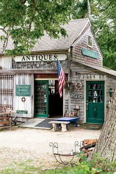 an antique shop with american flag on the front door and green shutters, sitting under a large tree