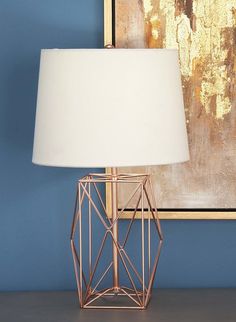 a table lamp with a white shade on it in front of a blue wall and painting