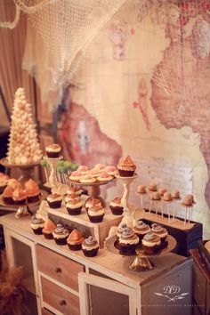 a table topped with lots of cupcakes next to a wall covered in maps