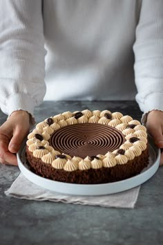 a person holding a chocolate cake with white frosting on it, in front of a plate