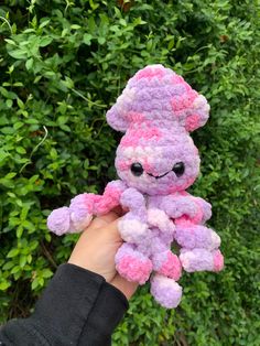 a hand holding a pink and purple teddy bear in front of some green bushes on a sunny day