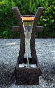 a water fountain in the shape of a cross on gravel ground with bushes behind it