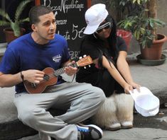 a man sitting on the ground playing a guitar next to a woman holding a paper bag