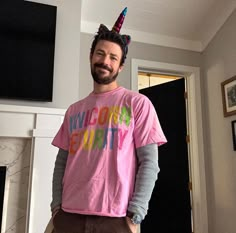 a man in a pink shirt with a unicorn horn on his head standing next to a fireplace