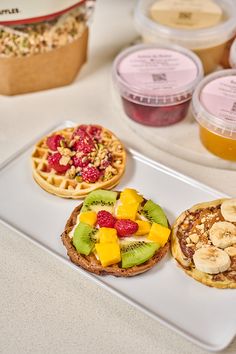 three waffles with fruit and nuts are on a white plate next to cups of yogurt