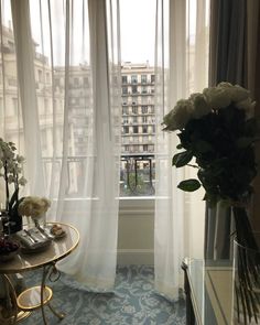 there is a vase with flowers on the table in front of the window that has sheer curtains