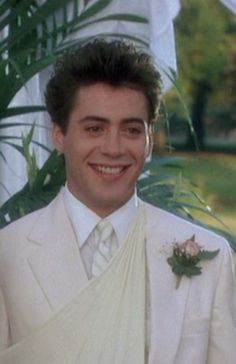 a man in a white suit and tie smiling at the camera with greenery behind him