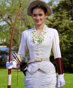 a woman in white dress and hat holding a bow and arrow while standing on grass