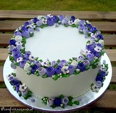 a white cake with purple and green flowers on it sitting on a wooden table in the grass