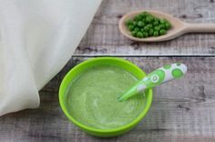 a green bowl with peas and a spoon in it on a wooden table next to a white cloth