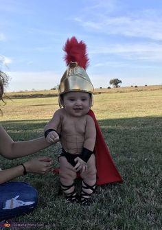 a woman kneeling down next to a baby dressed as a roman soldier with a red cape on it's head