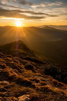 the sun is setting on top of a mountain with grass and rocks in the foreground