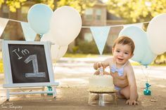 a baby sitting on the ground in front of a cake and balloons with a chalkboard sign