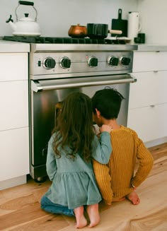 two children sitting on the floor in front of an oven looking at something inside it