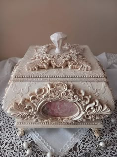 an ornate white box with a pink stone in the center on a lace doily