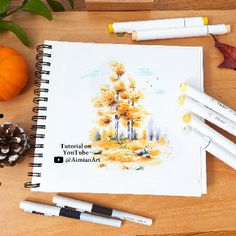 an open notebook sitting on top of a wooden table next to markers and pine cones