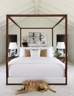 a dog laying on the floor in front of a bed with white sheets and pillows