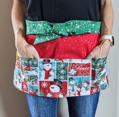 a woman wearing a christmas themed apron with her hands in her pockets and the belt undone