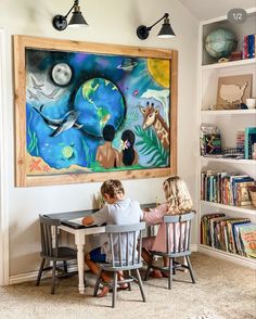 two children sitting at a table in front of a painting on the wall above them