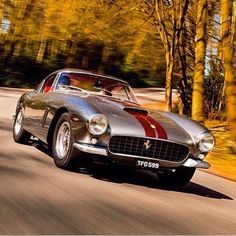 an old silver sports car driving down the road with trees in the backgroud