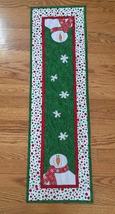 a green table runner with snowmen and polka dots on the border, sitting on a wooden floor