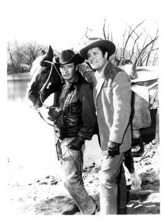 black and white photograph of two men standing next to each other with a horse in the background