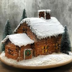 a gingerbread house with snow on the roof and trees around it, sitting on a plate