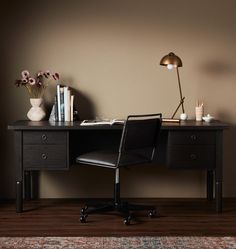 a desk with a chair, lamp and books on it in front of a brown wall