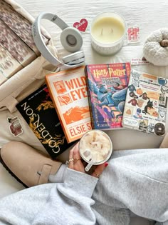 a person holding a cup of coffee next to books and headphones on a bed