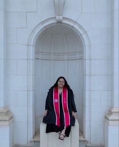 a woman sitting on top of a bench wearing a red and black graduation robe with her hands in her pockets