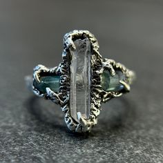 Forged from recycled sterling silver, this ring features a trio of stones--a central raw quartz crystal point flanked by two rough teal tourmaline stones. Skillfully sculpted from wax and cast using the revered lost wax casting technique, the ring possesses an ancient texture accentuated by a bewitching black patina. Beyond mere aesthetics, each stone is imbued with potent healing energies. The raw quartz crystal is a conduit for amplifying energy and clarity, while teal tourmaline is revered for its soothing properties and capacity to balance emotions. Sized at 8.5 and resizable upon request. Sterling silver, known for its healing properties, is believed to enhance the powers of the moon, promoting emotional balance and calming energies. Amethyst, with its rich violet hue, is thought to b Bridal Anklet, Mystical Jewelry, Alternative Engagement Ring, Raw Quartz Crystal, Raw Quartz, Tourmaline Stone, Antique Engagement, Alternative Engagement Rings, Antique Engagement Rings