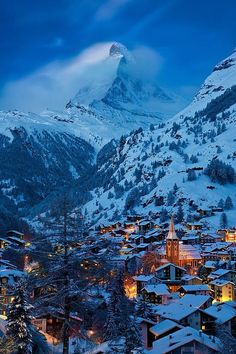 a snowy mountain with houses in the foreground and lit up buildings on the other side