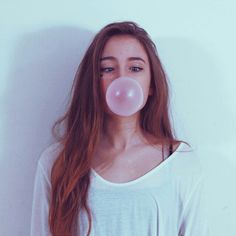 a woman with long red hair blowing a bubble into her mouth while standing in front of a white wall