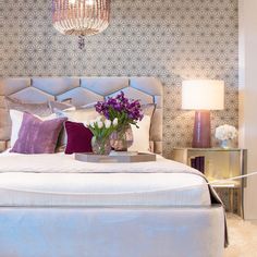 a bed with purple and white pillows on top of it next to a table with flowers